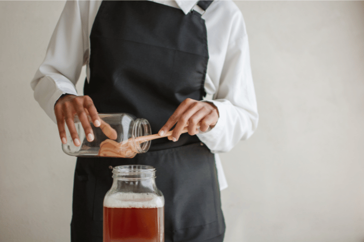 A woman pouring kombucha while holding the scoby with a wooden spoon