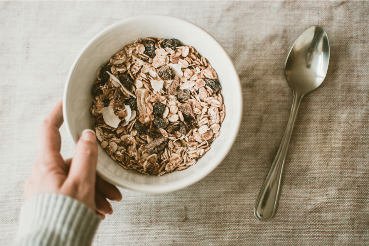 A hand holding a bowl of muesli (oats and raisins), above a linen placemat and silver spoon