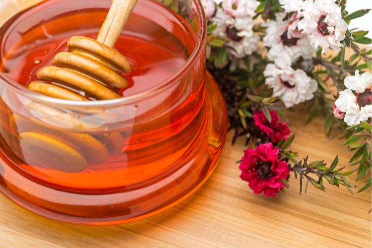 A glass jar full of Manuka Honey on a table, with Manuka flowers